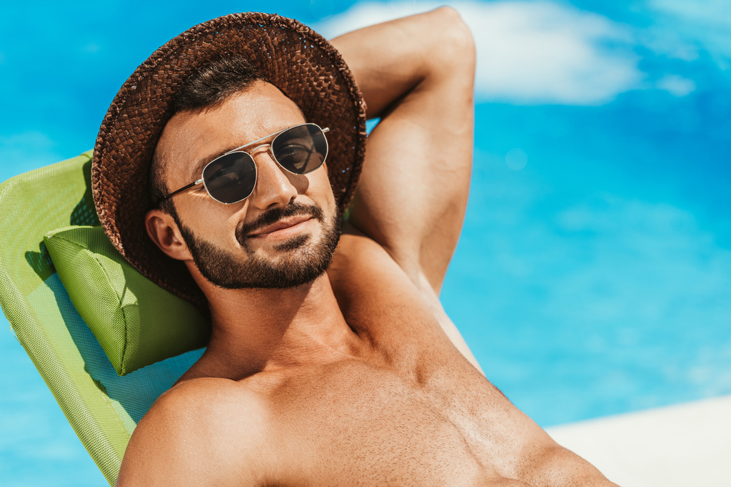 όμορφος άντρας με γυαλιά ηλίου και ψάθινο καπέλο χαλαρώνοντας στις ξαπλώστρες στη πισίνα - Φωτογραφία, εικόνα