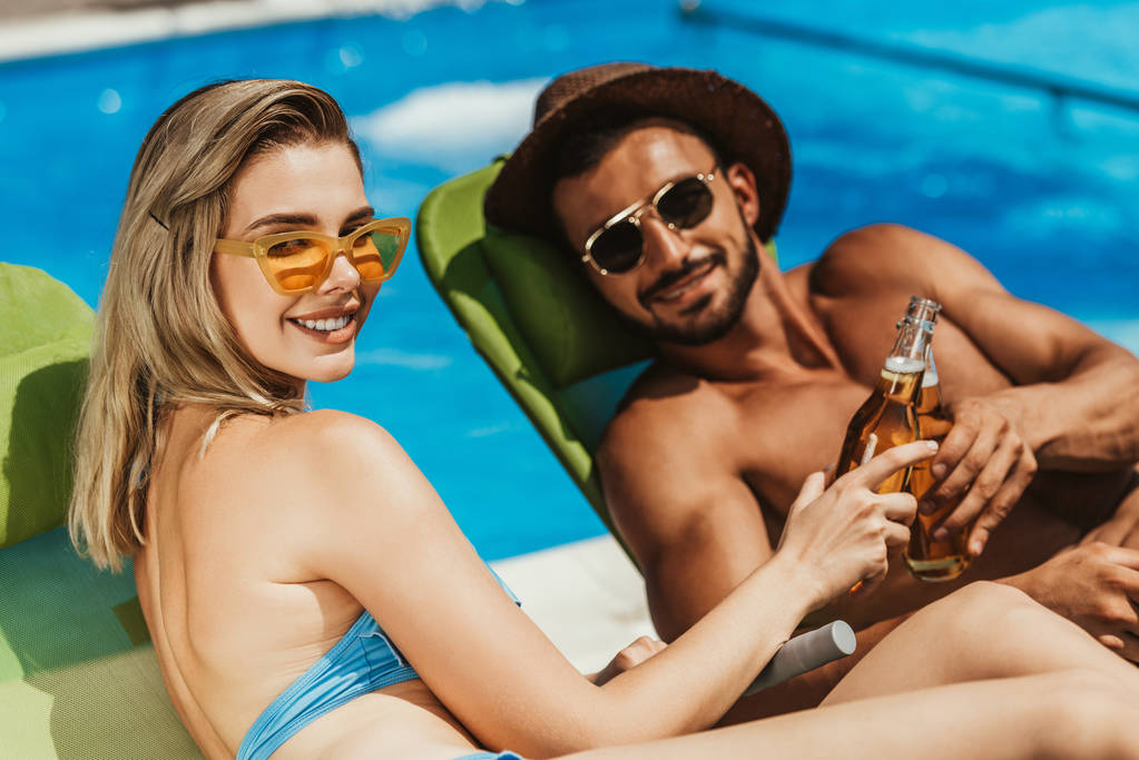 sonriente pareja en gafas de sol tintineo con botellas de cerveza mientras está acostado en tumbonas
 - Foto, Imagen