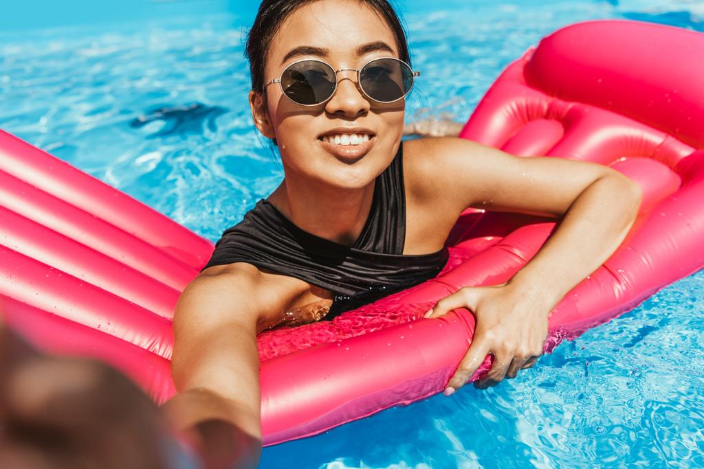 Uśmiechnięta dziewczyna azjatyckich w okulary biorąc selfie na nadmuchiwany materac w basenie - Zdjęcie, obraz