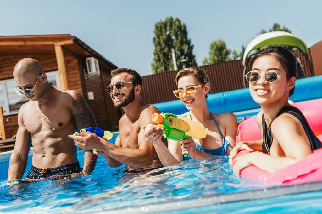 sonrientes amigos multiculturales divirtiéndose con pistolas de agua en la piscina
 - Foto, imagen