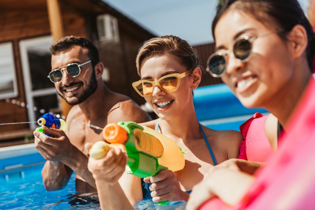pessoas multiétnicas em óculos de sol brincando com armas de água na piscina
 - Foto, Imagem
