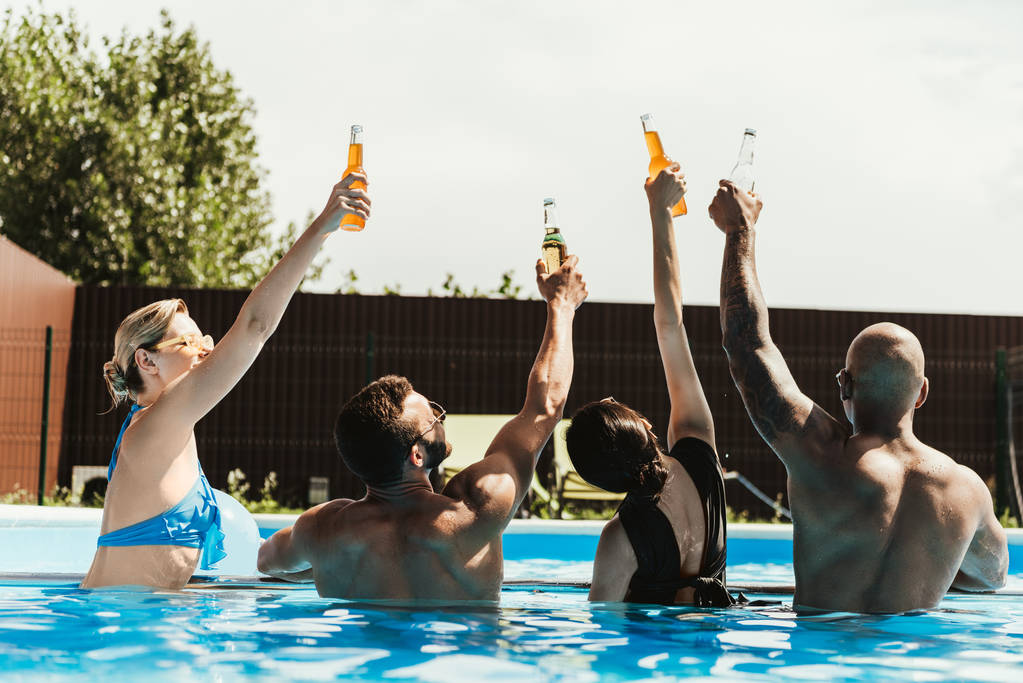 Rückansicht von multikulturellen Freunden, die im Schwimmbad Bierflaschen in die Höhe stemmen - Foto, Bild