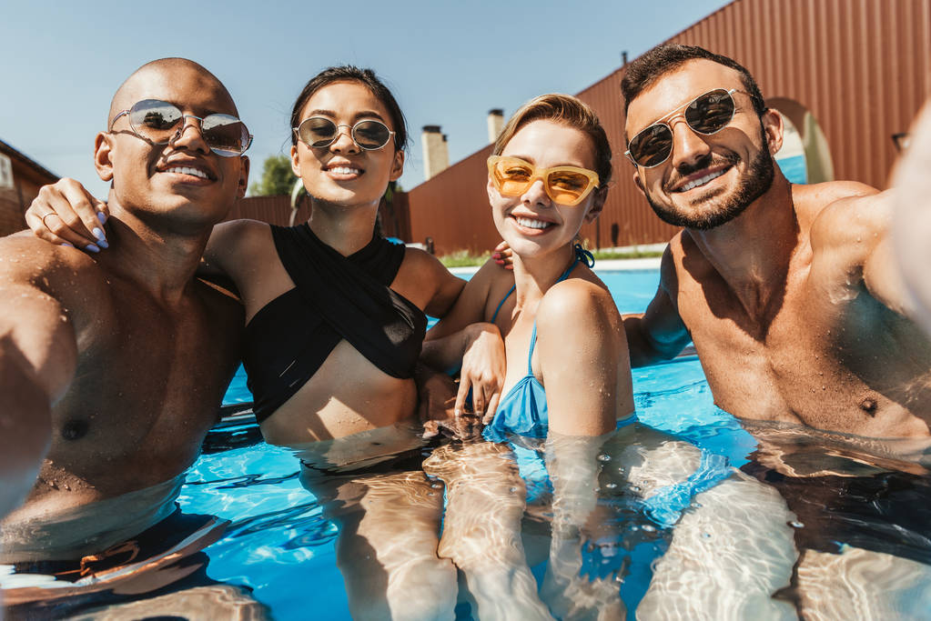happy multiethnic friends in swimsuits and sunglasses posing in swimming pool - Photo, Image