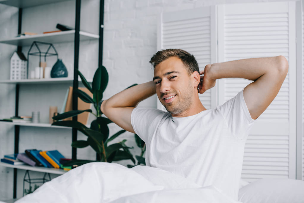 pohledný mladý muž protažení rukou a usmívá se na fotoaparátu zatímco sedí na posteli v ranní - Fotografie, Obrázek