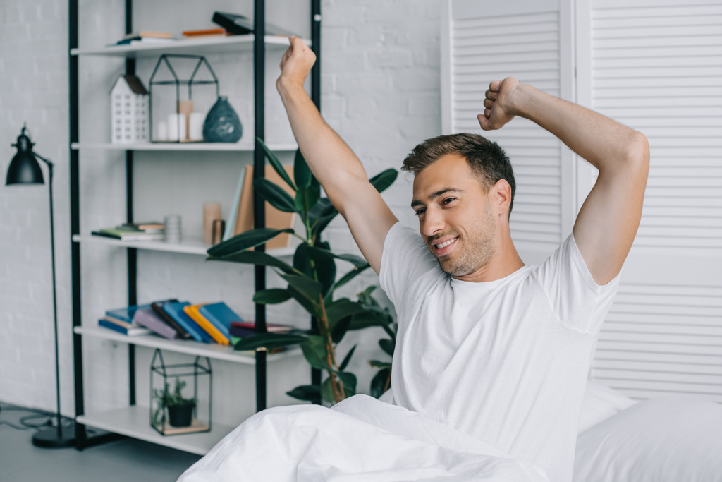 smiling young man stretching and looking away while sitting on bed - Photo, Image
