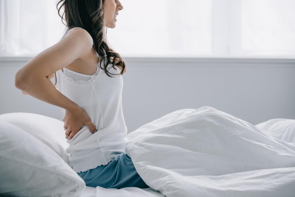 Schnappschuss einer jungen Frau im Schlafanzug, die unter Rückenschmerzen leidet, während sie im Bett sitzt  - Foto, Bild