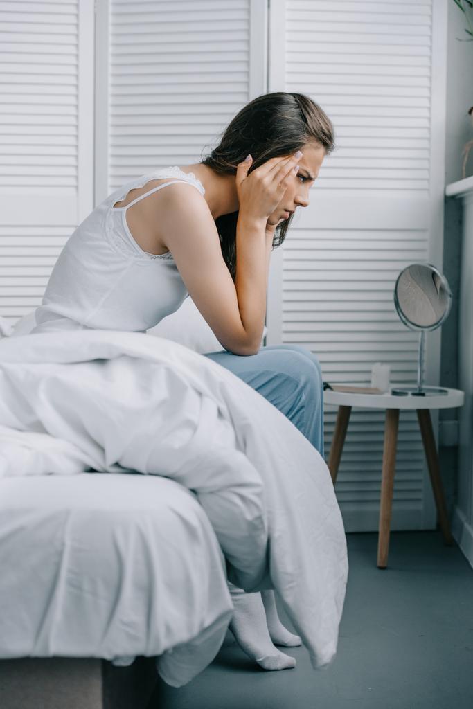 Zijaanzicht van de jonge vrouw in pyjama's lijden aan hoofdpijn op bed - Foto, afbeelding