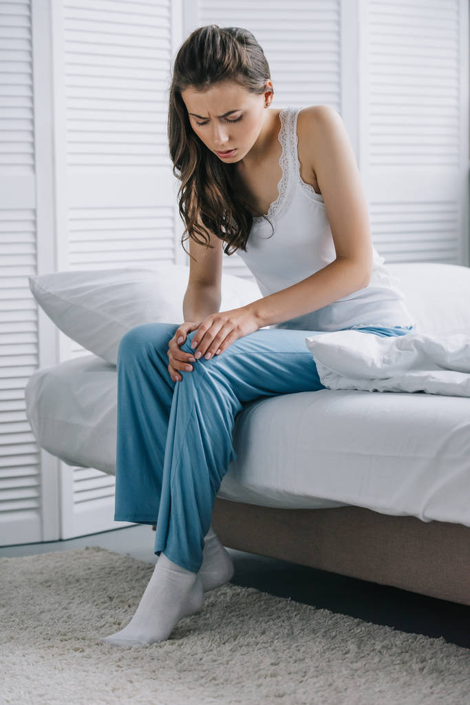 ελκυστική νεαρή γυναίκα στο πιτζάμες υποφέρουν από πόνο στο γόνατο, ενώ κάθεται στο κρεβάτι στο σπίτι - Φωτογραφία, εικόνα