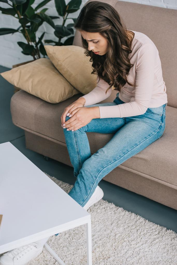 pohled z vysokého úhlu ženy s bolestí v koleně sedí na gauči doma - Fotografie, Obrázek