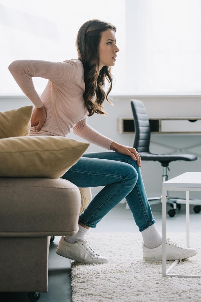 side view of young woman sitting on sofa and suffering from back pain  - Photo, Image