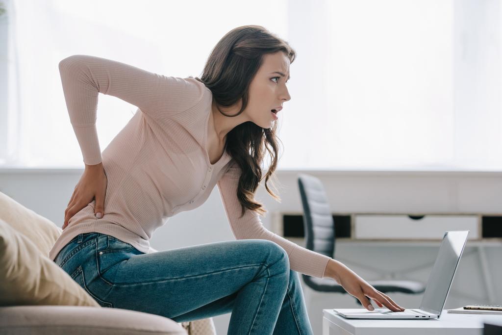 side view of young woman suffering from back pain while using laptop at home    - Photo, Image