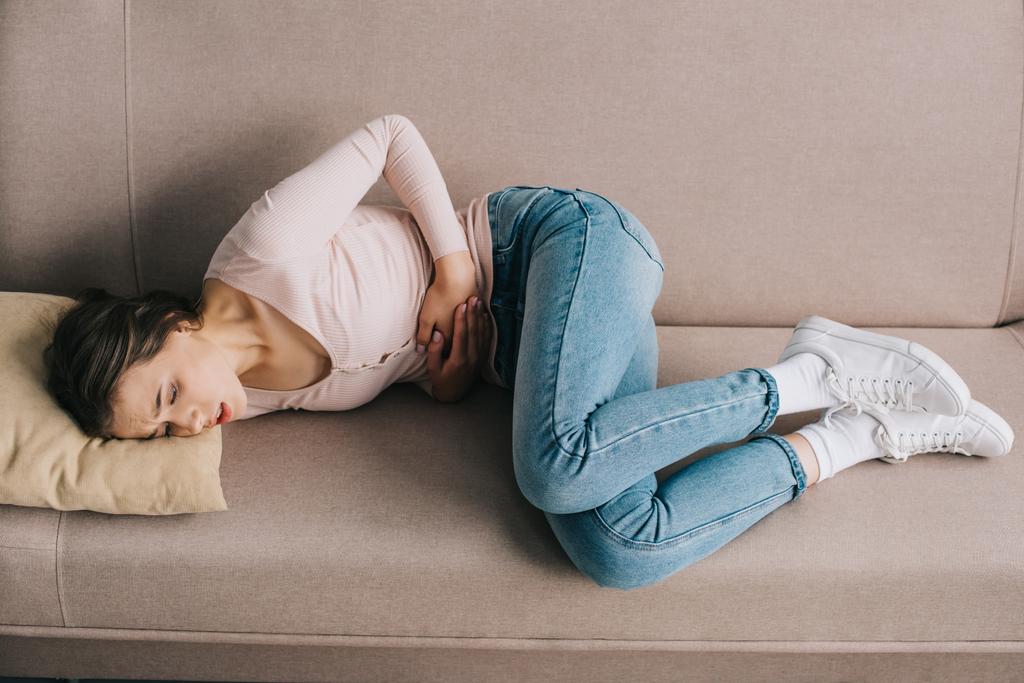 visão de alto ângulo da jovem mulher que sofre de dor de estômago enquanto deitada no sofá em casa
 - Foto, Imagem
