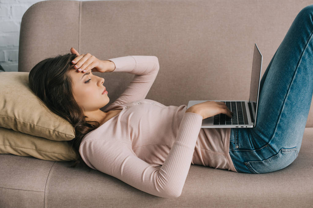 ソファの上に横たわっている間ノート パソコンを使用して頭痛を持つ若い女性の側面図 - 写真・画像