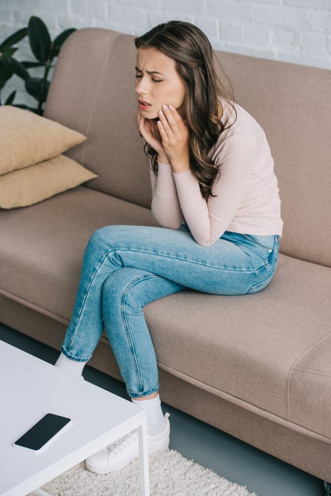 visão de alto ângulo da jovem mulher que sofre de dor de dente enquanto está sentada no sofá em casa
 - Foto, Imagem