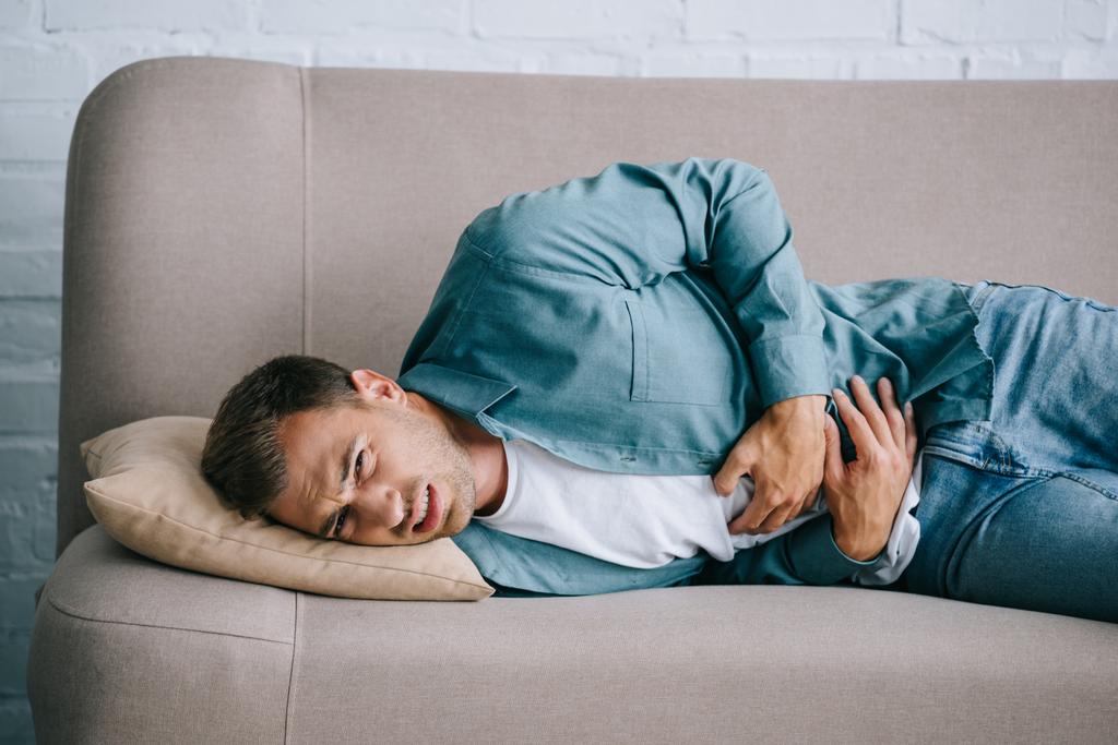 młody człowiek, leżąc na kanapie i cierpi na ból brzucha w domu  - Zdjęcie, obraz