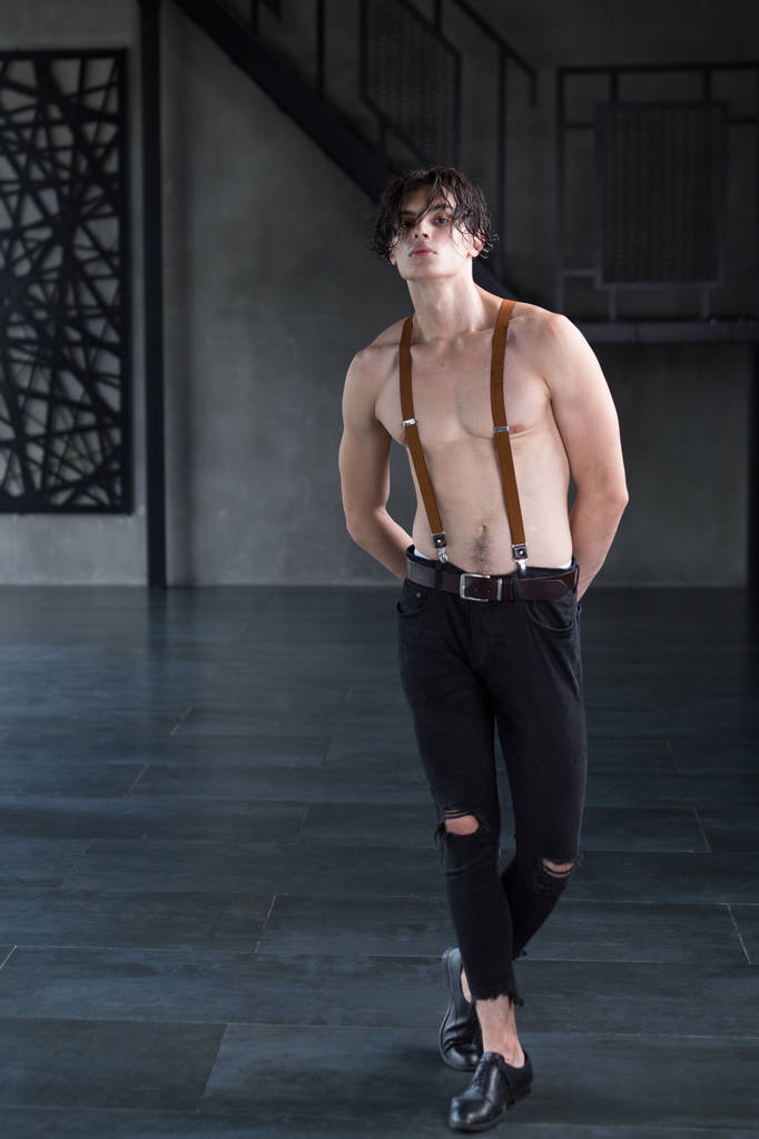 schöne hemdlose dunkelhaarige männliche Modell in zerrissenen Jeans und Hosenträgern in einem dunklen Raum - Foto, Bild