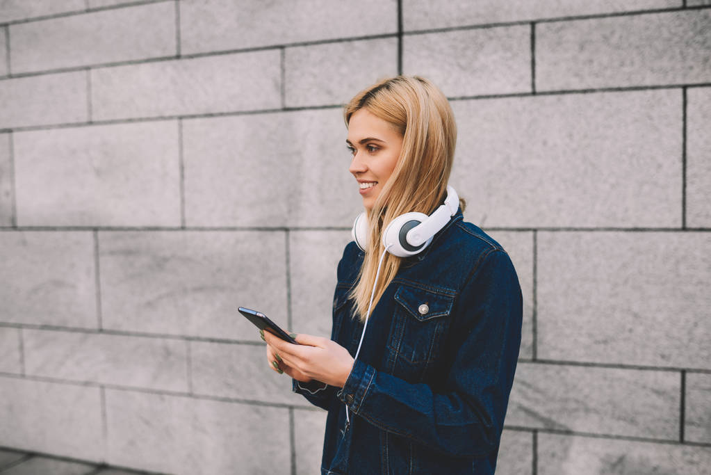 Menina hipster loira alegre com fones de ouvido modernos rindo durante as mensagens on-line no smartphone em pé sobre fundo promocional cinzento.Jovem positiva verificando e-mail no dispositivo de telefone
 - Foto, Imagem