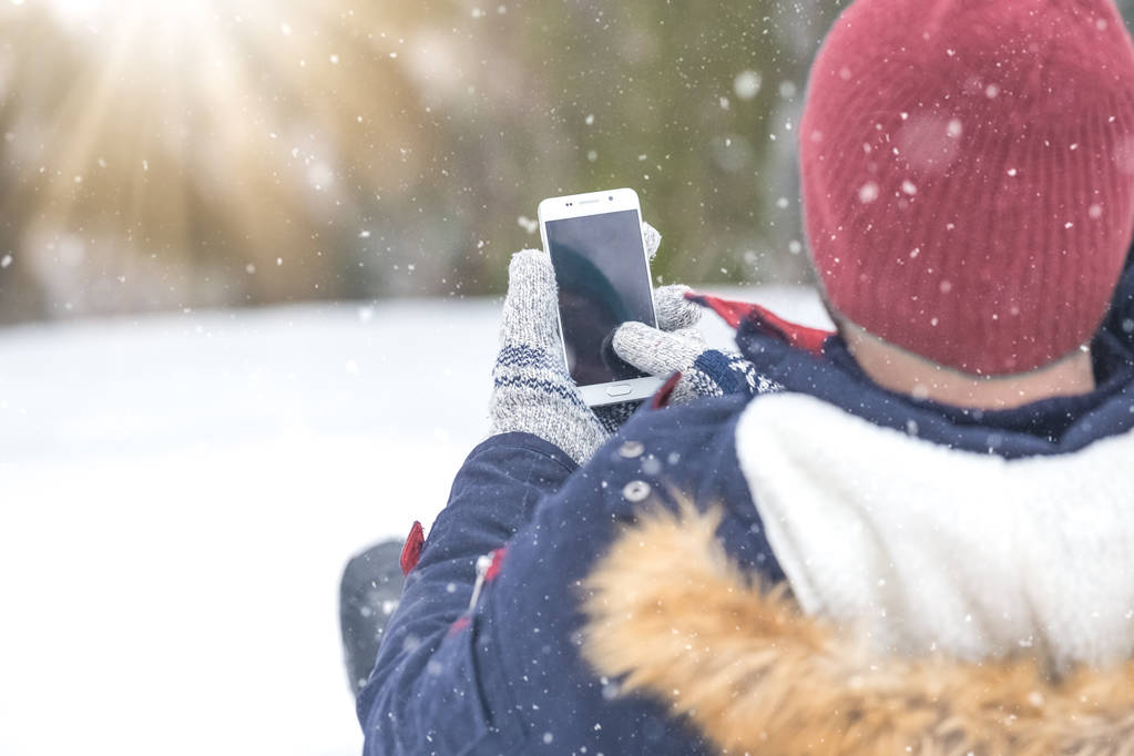 Πίσω όψη του ανθρώπου κάθεται στο χιόνι και κρατώντας κενής οθόνης έξυπνο τηλέφωνο. - Φωτογραφία, εικόνα