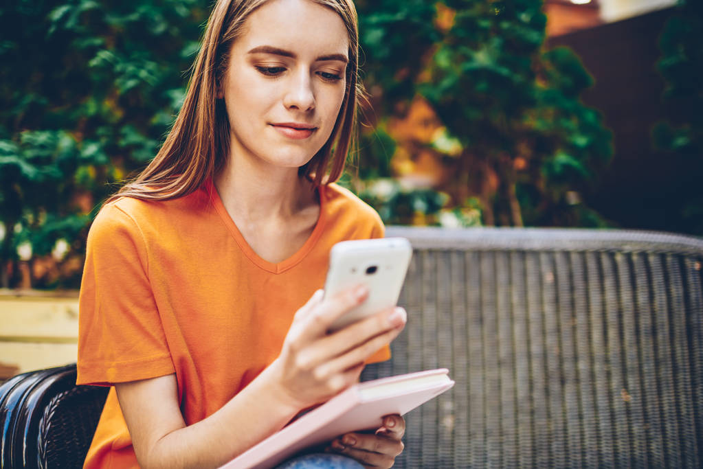 abgeschnittenes Bild der schönen Studentin beim Hochladen einer Anwendung und dem Senden von SMS in sozialen Netzwerken auf dem Handy, während sie sich auf einer Bank mit Lieblingsbuch ausruht. - Foto, Bild