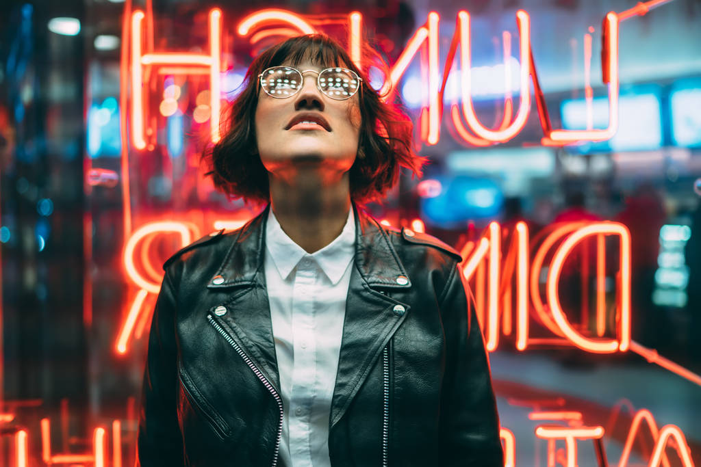 Κομψή γυναίκα μελαχρινή σε μοντέρνα ενδυμασία και γυαλιά κοιτώντας ψηλά απολαμβάνοντας τη νυχτερινή ζωή στην πόλη. Πανέμορφο hipster κορίτσι ντυμένο με Δερμάτινη ζακέτα στέκεται έξω στο δρόμο με φωτισμό νέον πόλη - Φωτογραφία, εικόνα