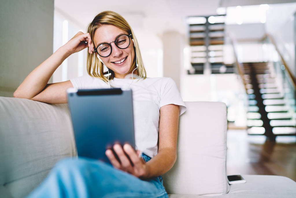 Optik gözlükler komik eğitsel bir dokunmatik yüzey evde kanepede oturan izlerken pozitif sarışın genç kadın. Kulaklık takipçileri Tablet yorum okuma sırasında gülüyor içinde neşeli blogger - Fotoğraf, Görsel