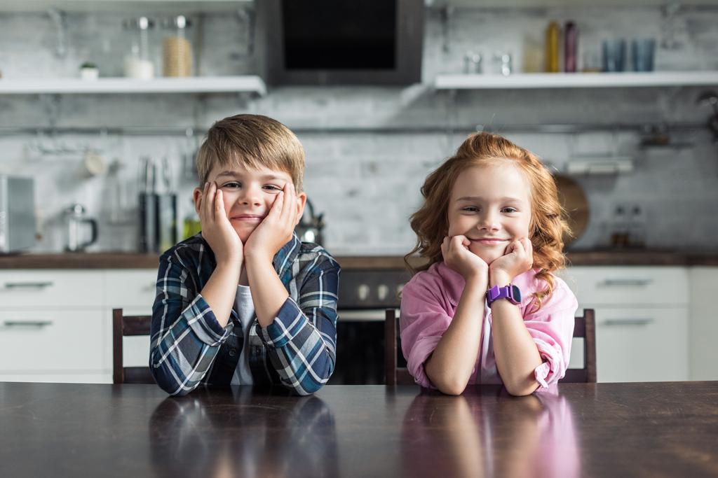 usmívající se malý bratr a sestra seděli v kuchyni a při pohledu na fotoaparát - Fotografie, Obrázek