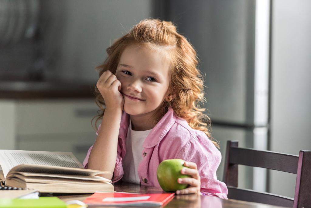 Nettes kleines Schulmädchen mit grünem Apfel schaut während der Hausaufgaben in die Kamera - Foto, Bild