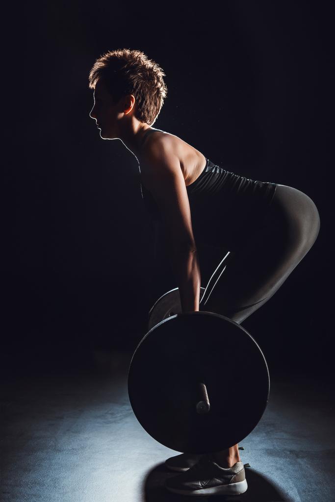 вид сбоку на спортсменку, делающую упражнения с штангой в тренажерном зале, черный фон
 - Фото, изображение