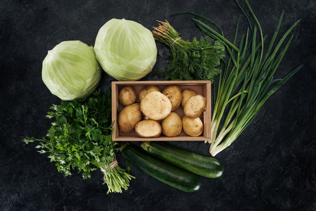 το Top view πατάτες σε κουτί με τοποθετημένα τα πράσινα λαχανικά σε μαύρο μαρμάρινη επιφάνεια - Φωτογραφία, εικόνα