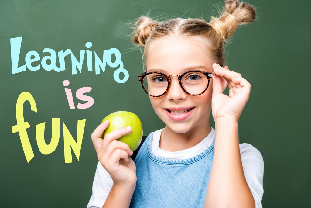 Schulkind hält Apfel in der Nähe der Tafel und berührt Gläser mit dem Schriftzug "Lernen macht Spaß" - Foto, Bild