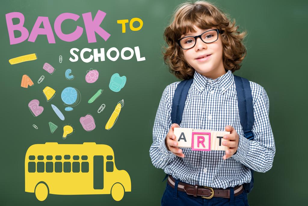 schoolboy holding wooden cubes with word art near blackboard, with icons, bus and "back to school" lettering - Photo, Image