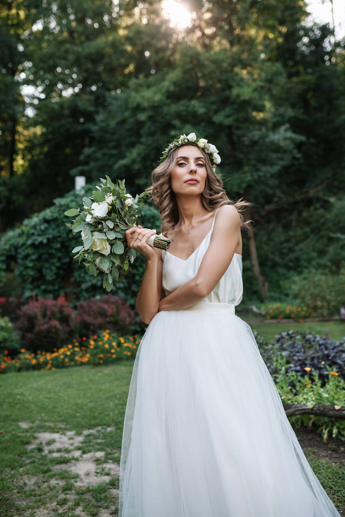 屋外の花束と花輪のエレガントな白いドレスで美しい金髪の幸せな花嫁 - 写真・画像