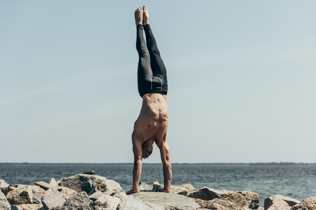sporty shirtless man doing handstand (adho mukha vrksasana) on rocky seashore - Photo, Image
