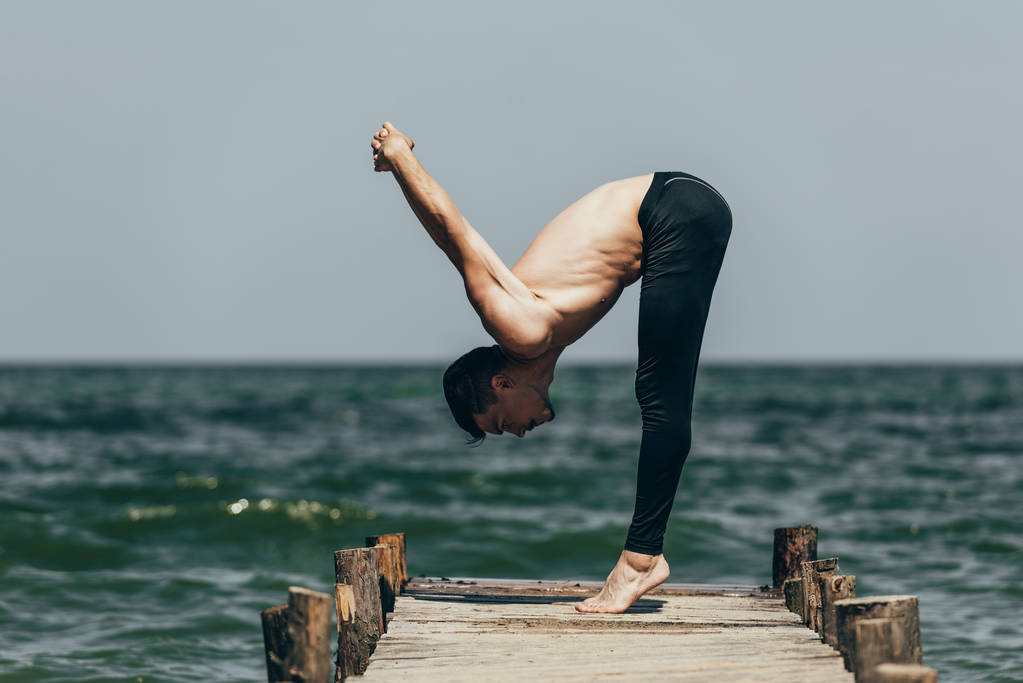 Seitenansicht eines hemdlosen Mannes, der Yoga in uttanasana-Pose auf einem Pier praktiziert - Foto, Bild