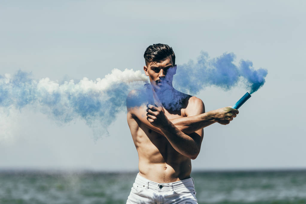 homme torse nu musclé avec des bâtons de fumée bleus et blancs devant l'océan
 - Photo, image