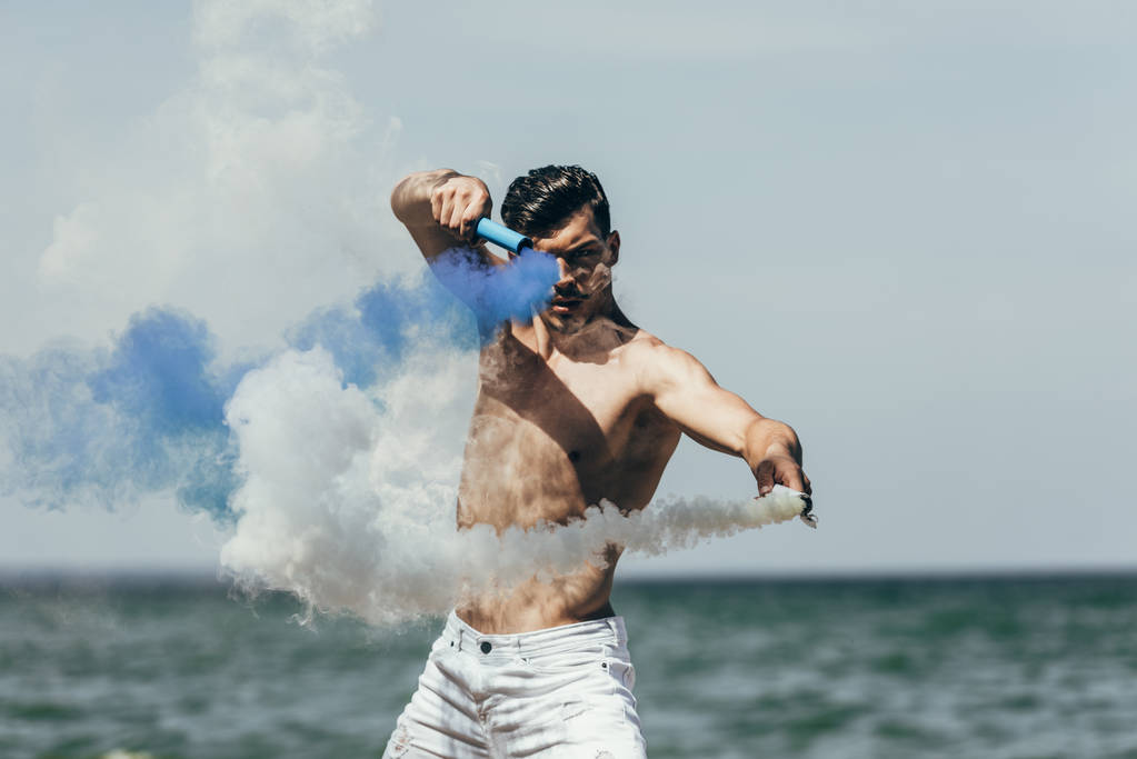 海の前に青と白の煙棒踊り魅力的な上半身裸の男 - 写真・画像