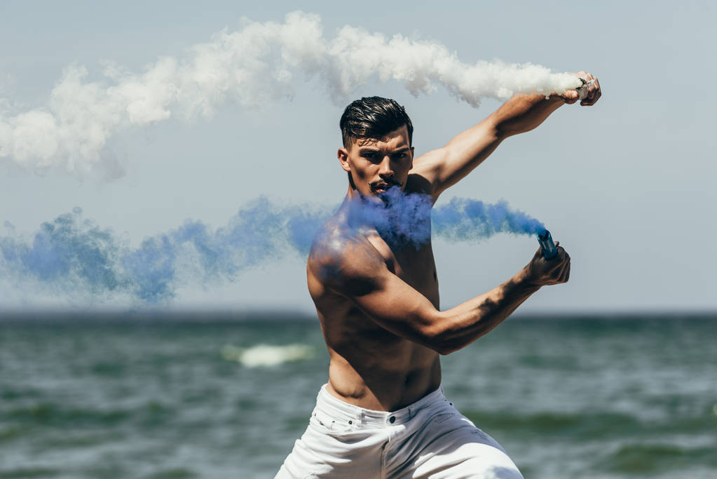 bonito sem camisa homem dançando com azul e branco varas de fumaça na frente do oceano
 - Foto, Imagem