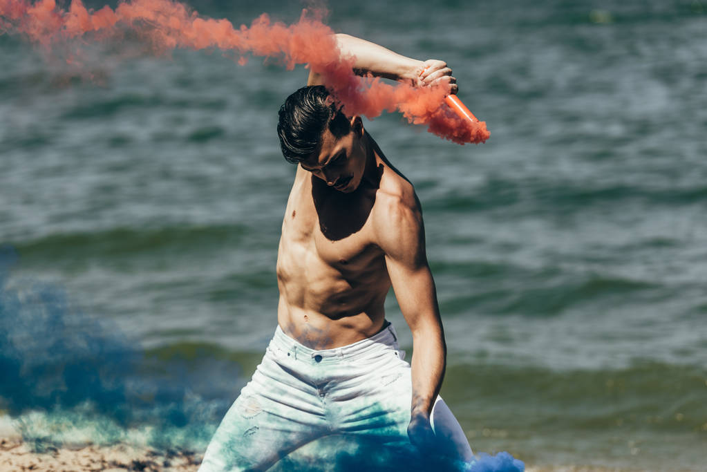 homme torse nu dansant avec des bâtons de fumée rouges et bleus devant la surface de l'eau
 - Photo, image