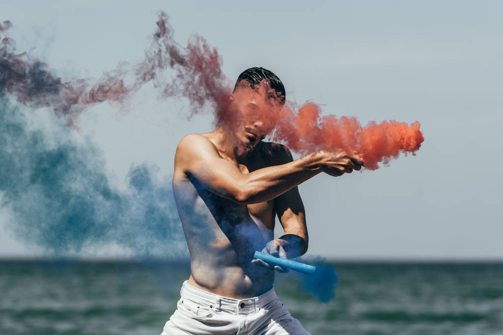 homem sem camisa atraente dançando com varas de fumaça vermelhas e azuis na frente da vista do oceano
 - Foto, Imagem