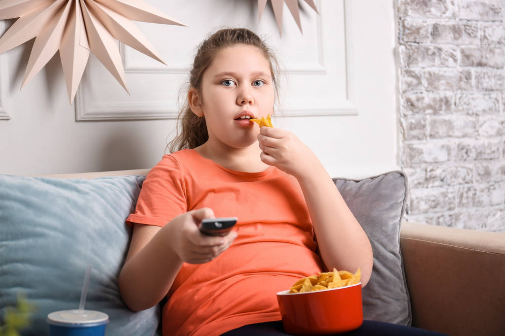 Дівчина з надмірною вагою дивиться телевізор із закусками в приміщенні
 - Фото, зображення