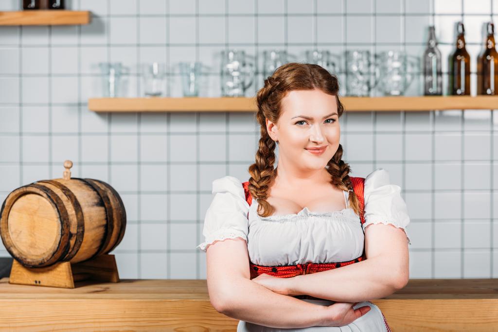 улыбающаяся Октоберфест официантка в традиционном баварском платье со скрещенными руками, стоящая возле барной стойки с пивной дубинкой
  - Фото, изображение