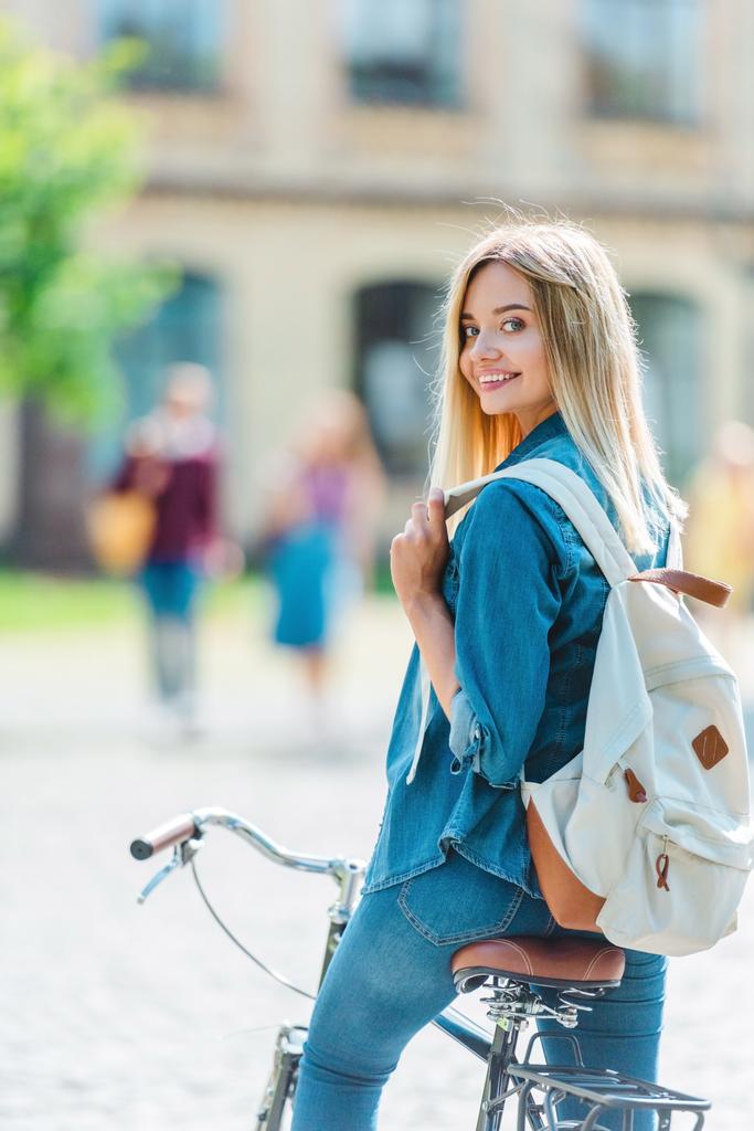 valikoiva keskittyä hymyilevä nuori opiskelija reppu polkupyörän katselee kameraa kadulla
 - Valokuva, kuva