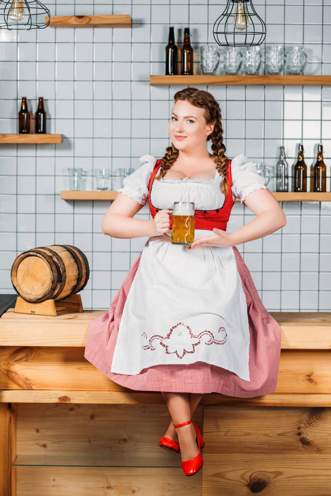 attractive oktoberfest waitress in traditional bavarian dress showing mug of light beer while sitting on bar counter - Photo, Image