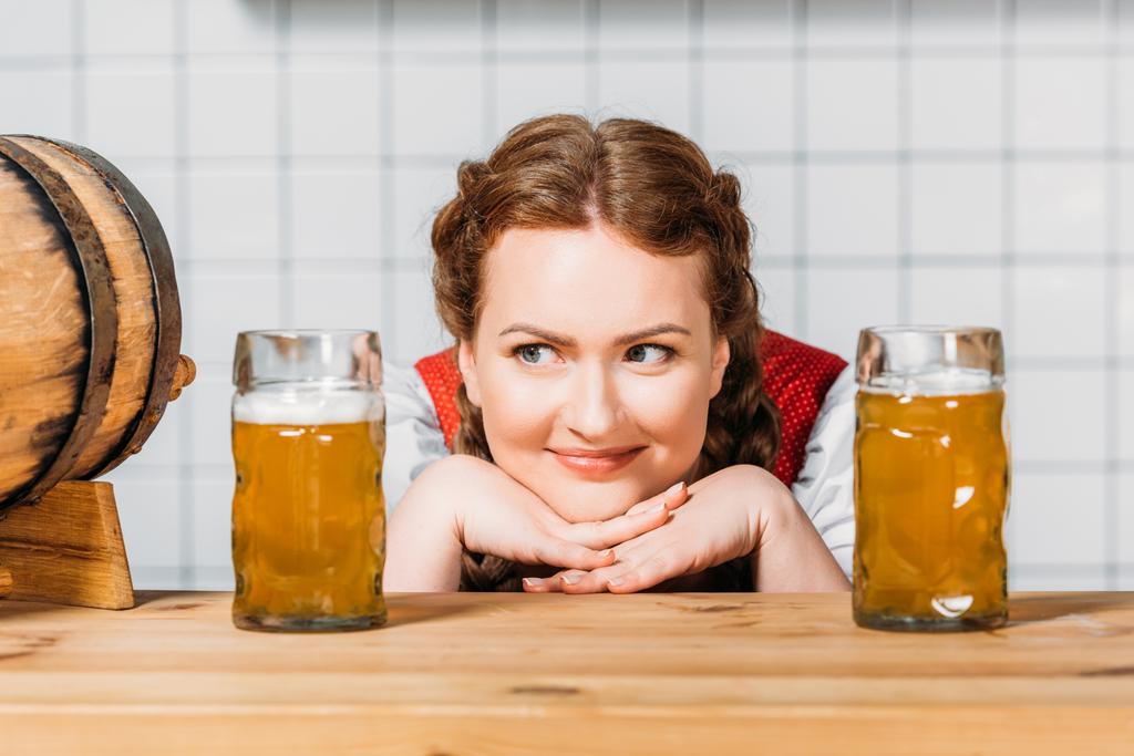 sorrindo oktoberfest garçonete em vestido tradicional bávaro sentado entre duas canecas de cerveja leve no balcão do bar
 - Foto, Imagem