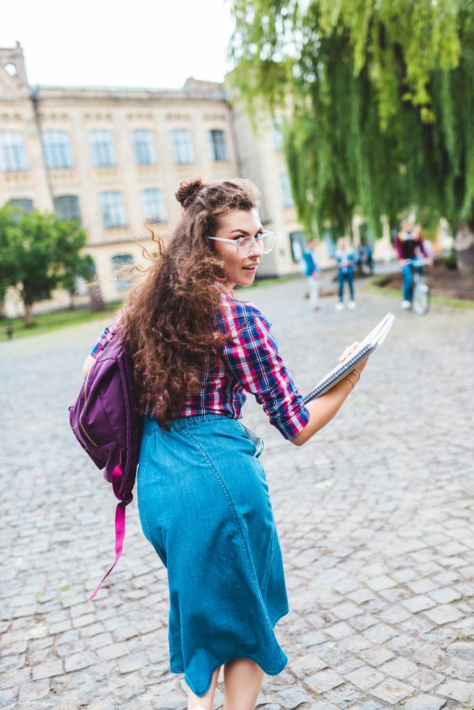 Hátulnézet a fiatal diák a szemüvegek, a hátizsák és notebook-ok, futó utca - Fotó, kép