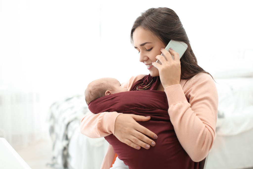 Νεαρή γυναίκα με το μωρό σε σφεντόνα μιλάμε για κινητό τηλέφωνο σε εσωτερικούς χώρους - Φωτογραφία, εικόνα