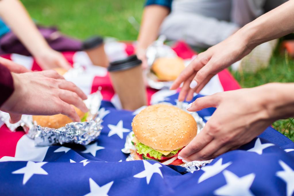částečný pohled přátel s hamburgery a americká vlajka na zelené trávě v parku - Fotografie, Obrázek