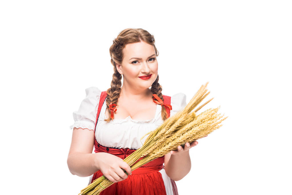 lachende oktoberfest serveerster in traditionele Beierse kleding houden van tarwe oren geïsoleerd op witte achtergrond - Foto, afbeelding