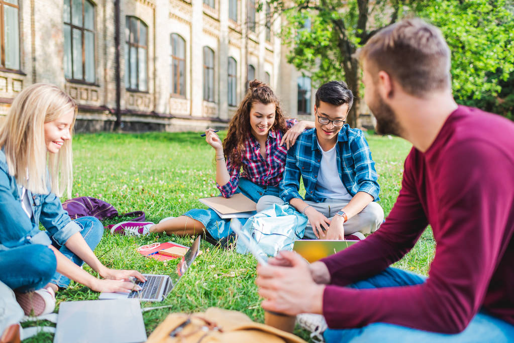 πολυφυλετικές χαμογελώντας φοιτητές με ψηφιακές συσκευές κάθεται στο πράσινο γρασίδι στο πάρκο - Φωτογραφία, εικόνα