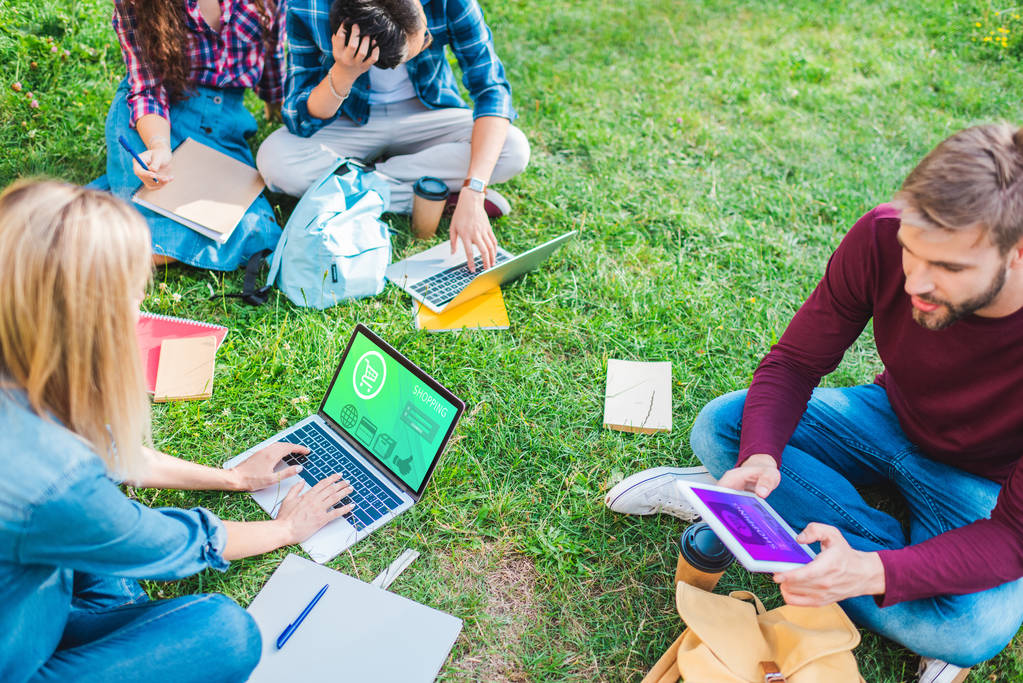 частковий вид на багатоетнічних студентів з блокнотами та цифровими пристроями, що сидять на зеленій траві в парку
 - Фото, зображення